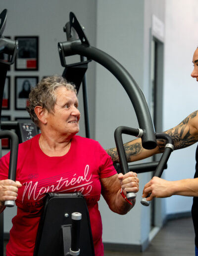 Une dame âgée se fait coacher par une coach sportif Olympe Fitness sur un appareil de musculation